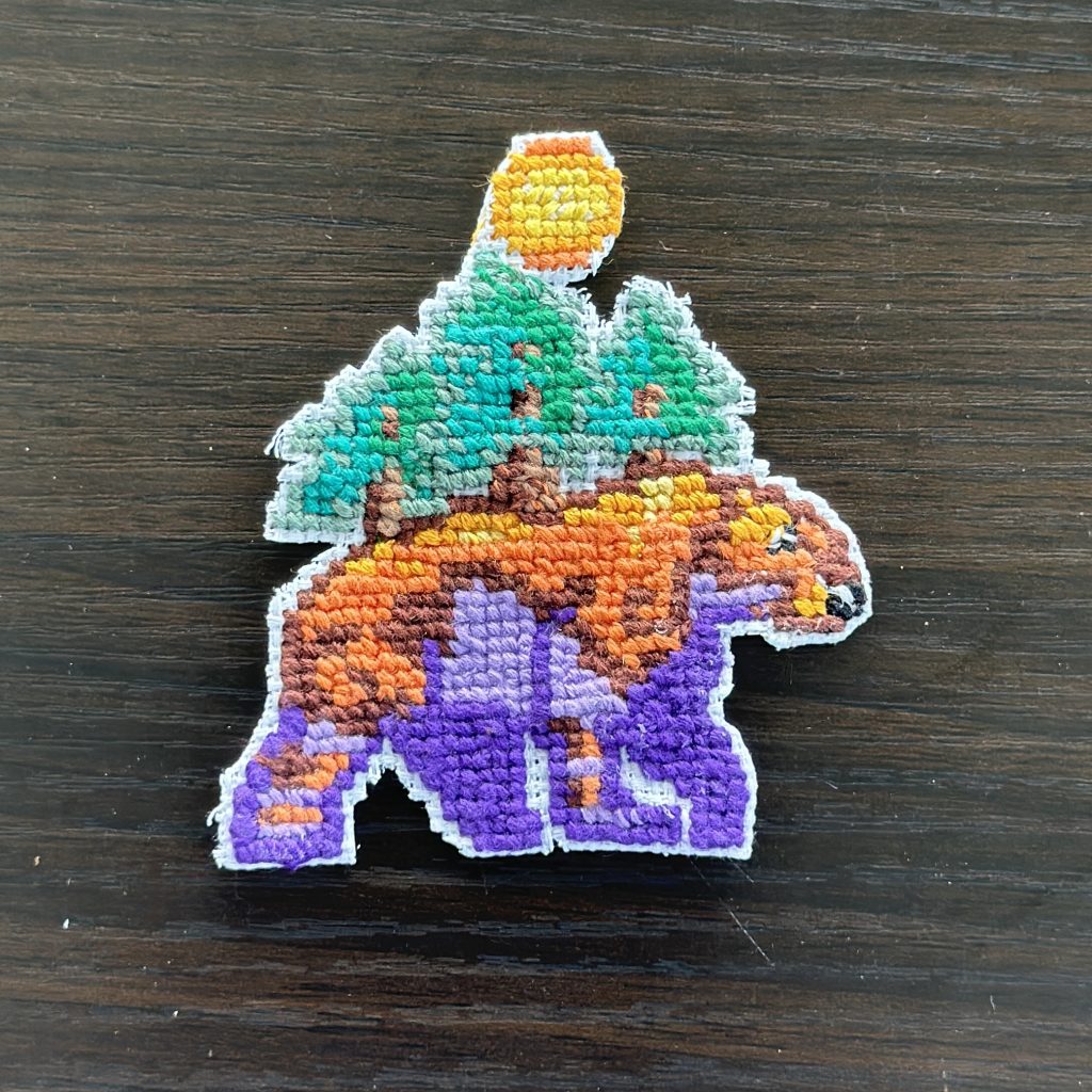 Cross-stitched patch of bear in front of deciduous trees under a full sun in vibrant purple, orange, and green tones.
