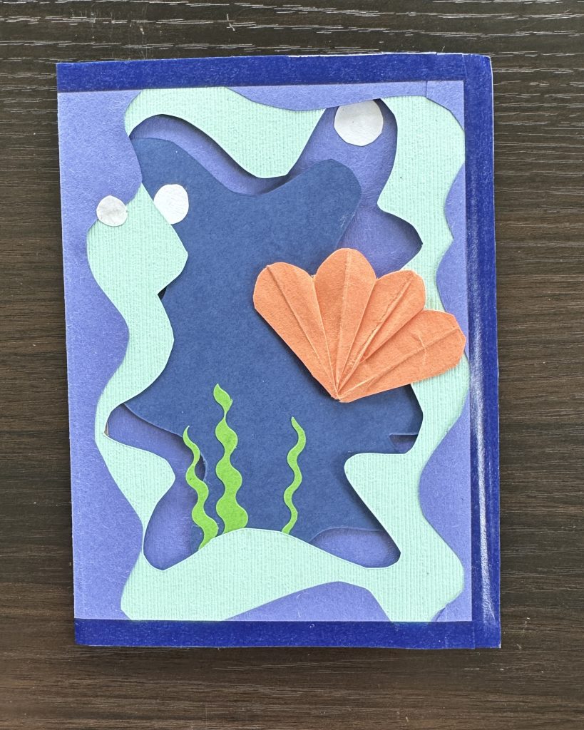 Card displaying an underwater scene with layered water cutouts, origami shell, seaweed, and bubbles.